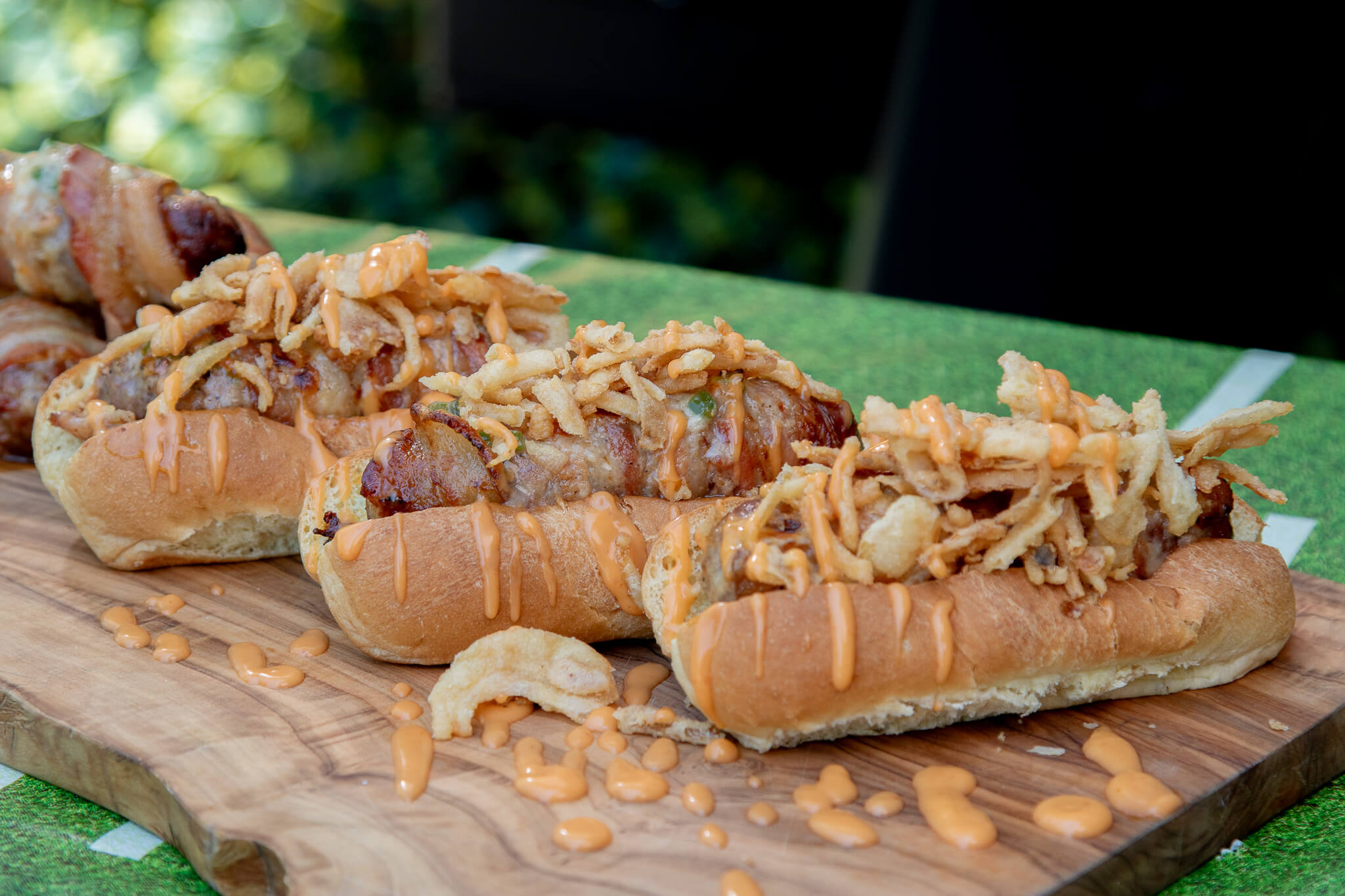 grilled sausages wrapped in bacon and topped with fried onions and spicy mayo on wooden cutting board